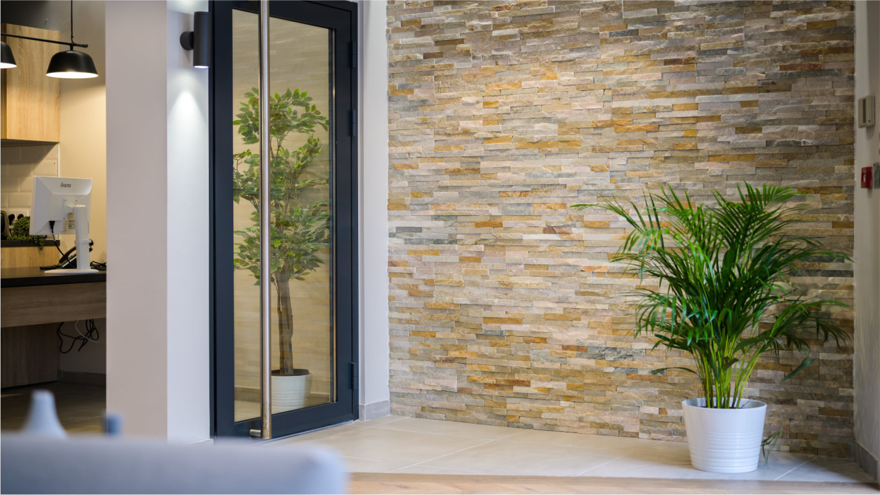 Alt text: A modern interior space featuring a textured stone wall in shades of beige and gray. To the right, a tall green plant in a white pot adds a touch of nature. In the background, a glass door leads to another area, with a sleek desk and monitor visible. The overall ambiance is bright and inviting.